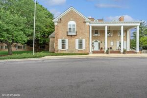 Two-story brick building with white columns, shutters, and balcony, set on a paved area with trees and lawn in the background.