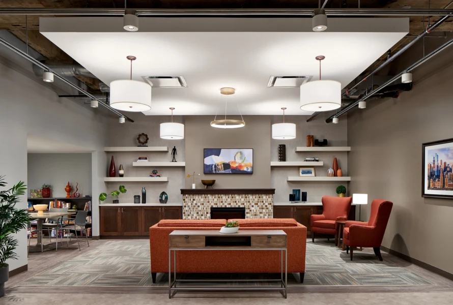Modern lounge area with orange seating, a central coffee table, built-in shelves, wall art, and overhead lighting, featuring a fireplace and two armchairs near the back wall.