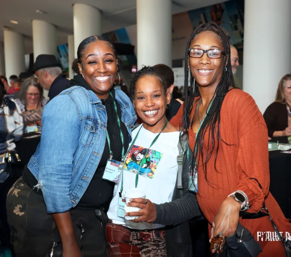 Three women smile and pose together at an indoor event; other attendees are visible in the background.