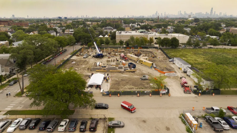 A construction site with cranes and vehicles is shown in a suburban area, surrounded by trees, parked cars, and residential buildings, with a city skyline in the distance.