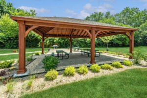 A wooden pavilion with a peaked roof shelters several picnic tables on a concrete patio, surrounded by green grass, shrubs, and trees under a blue sky.