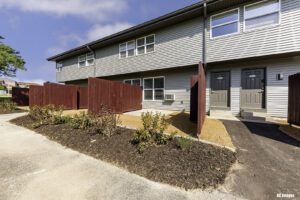 Two-story gray townhouse with two front doors, red privacy fences, brown mulch, small plants, and a concrete walkway under a partly cloudy sky.