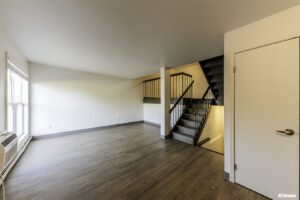 Unfurnished living room with wood flooring, white walls, large window, and a staircase with black railing leading upstairs.