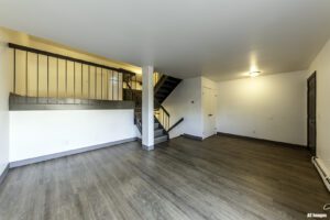 Unfurnished room with wood flooring, white walls, staircase with metal railing, and two doors. Ceiling light fixture is on.