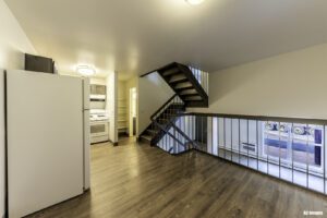 Apartment interior with wood flooring, an open kitchen with white appliances, a staircase with metal railings, and a large window facing the street.