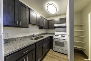 Modern kitchen with dark cabinets, granite countertops, stainless steel double sink, gas stove, range hood, wood flooring, and built-in pantry shelves on the right.