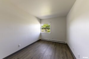 Empty room with white walls, a window with a view of trees, wood-look flooring, baseboard heater, and electrical outlets.
