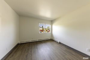 Empty room with white walls, wood-style flooring, baseboard heater, and a window showing buildings and trees outside.