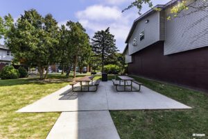 Two picnic tables on a concrete patio next to a building, surrounded by grass, trees, and clear sky on a sunny day.
