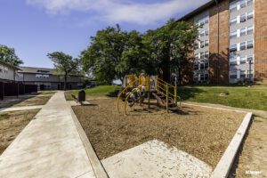 Playground with slides and climbing equipment on mulch, next to a sidewalk and multi-story brick apartment building under a blue sky.