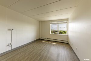 Empty room with light-colored walls, wood-patterned floor, a large window with outdoor view, and baseboard heating along the wall.
