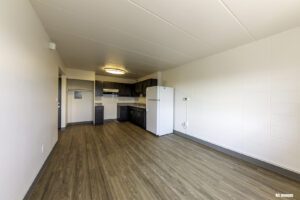 Unfurnished apartment interior with wood-style flooring, white walls, a small kitchen area with dark cabinets, a white refrigerator, and an overhead light fixture.
