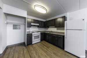 Modern kitchen with dark cabinets, white gas stove, refrigerator, and granite countertops. Open closet and wood-style flooring are visible. Bright ceiling light illuminates the space.