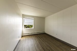 Empty room with white walls, a large window, baseboard heaters, and wood-look flooring. Sunlight enters through the window, illuminating the space.