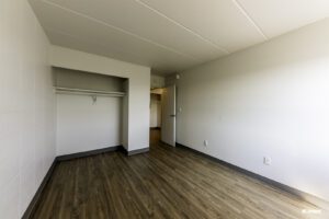 Empty room with light-colored walls, wood flooring, an open closet, electrical outlets, and a door leading to another space with similar flooring.
