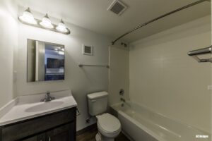 Modern bathroom with a vanity sink, mirror, toilet, and bathtub with shower, featuring white walls and wood-style flooring.
