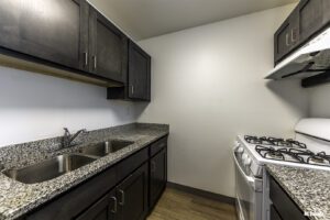 Narrow kitchen with dark cabinets, granite countertops, double sink, gas stove, range hood, and wood flooring.