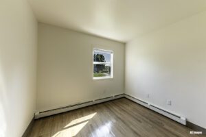 Empty room with light-colored walls, a window letting in natural light, wood-style flooring, and baseboard heaters along two walls.