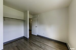 Empty room with wood flooring, open closet with shelf and hanging rod, white walls, baseboard heater, and an open door leading to a hallway with shelves.