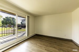 Empty room with wood flooring, white walls, and a large sliding glass door opening to a balcony overlooking a green outdoor area with trees and a building in the background.