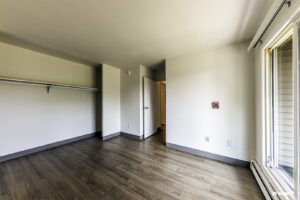 Empty room with hardwood flooring, open closet with shelf and rod, white walls, a door, and a large window letting in natural light.