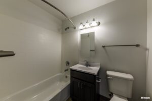 Bathroom with a white tub-shower combo, dark vanity with sink and mirror, toilet, wall light fixture, and towel bars.