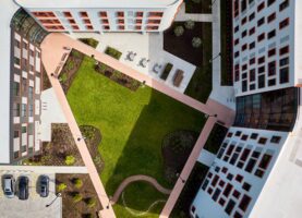 Aerial view of a modern building complex with three structures forming a triangular courtyard, featuring grass, pathways, seating areas, and parked cars.