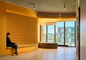 A person sits on a built-in yellow bench using a phone in a modern, yellow-walled office space with large windows and city views.