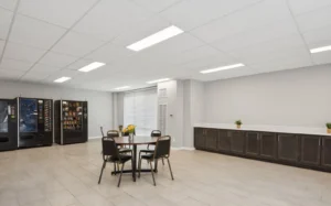 A break room with vending machines, a round table with four chairs, long countertop with cabinets, and neutral-colored walls and floor.