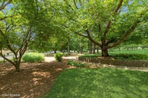 A sunny park area with lush green trees, grass, a stone retaining wall, and a path leading past a black metal fence.