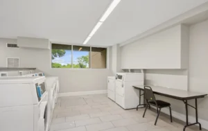 A clean, bright laundry room with several white washing machines and dryers, a black chair, a folding table, tiled floor, and a window showing trees outside.