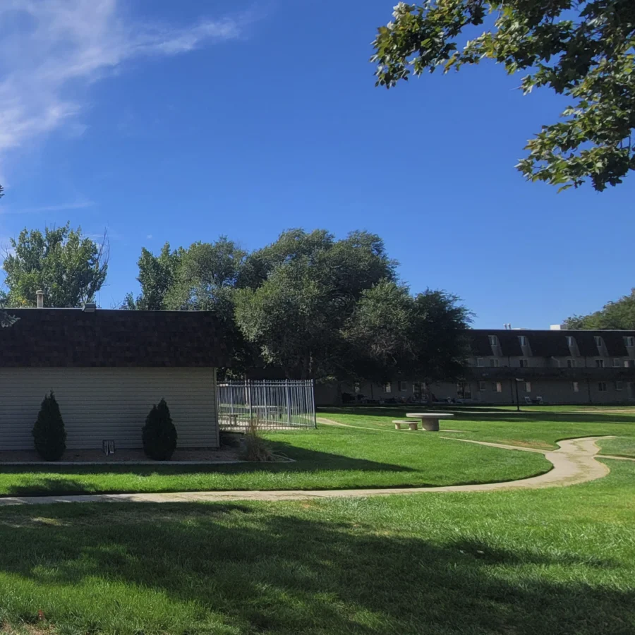 A well-maintained grassy courtyard with winding sidewalks, a picnic table, trees, a small building, and an apartment building in the background under a clear blue sky.