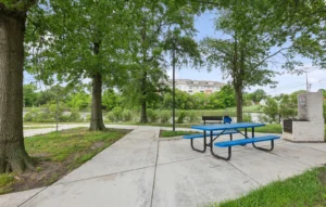 A blue picnic table sits on a concrete patio under trees near a walking path, with a bench, grill, and building visible in the background.