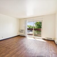 Unfurnished living room with wood flooring, white walls, a wall-mounted air conditioner, and sliding glass doors leading to a balcony with a view of trees.