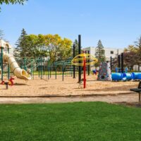 Outdoor playground with various equipment including a slide, climbing structures, blue tunnels, and monkey bars, set on sand with grass in the foreground and trees in the background.