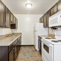 A narrow kitchen with dark cabinets, light countertops, white appliances including a fridge, stove, and microwave, and a single overhead light fixture.