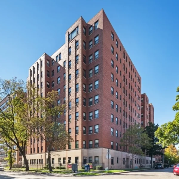 A tall brick apartment building with multiple windows, situated on a street corner with trees and a clear blue sky.