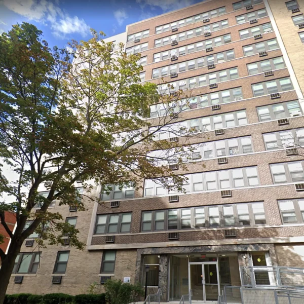 A tall, beige brick apartment building with many windows and air conditioning units, a tree in front, and a glass entrance at street level.