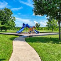 A paved path leads to a playground with blue slides and climbing equipment, surrounded by grass, trees, and a clear blue sky.