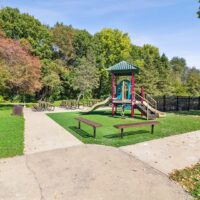 A small playground with a slide and climbing structure, surrounded by grass and trees, with picnic tables and benches nearby on a sunny day.