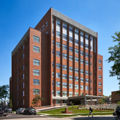 A multi-story red brick building with large windows and a covered entrance, surrounded by a lawn and parked cars under a clear blue sky.