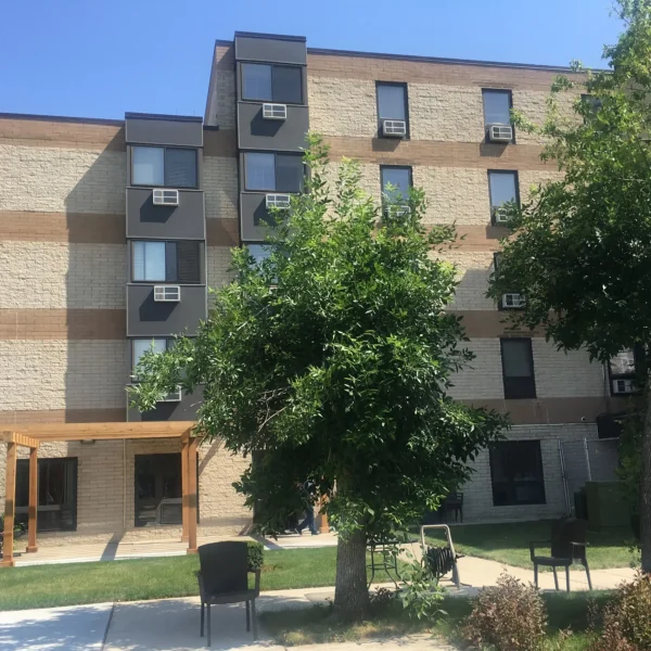 Three-story apartment building with beige and brown brick exterior, window air conditioners, and a small garden area with trees, chairs, and a pergola in front.
