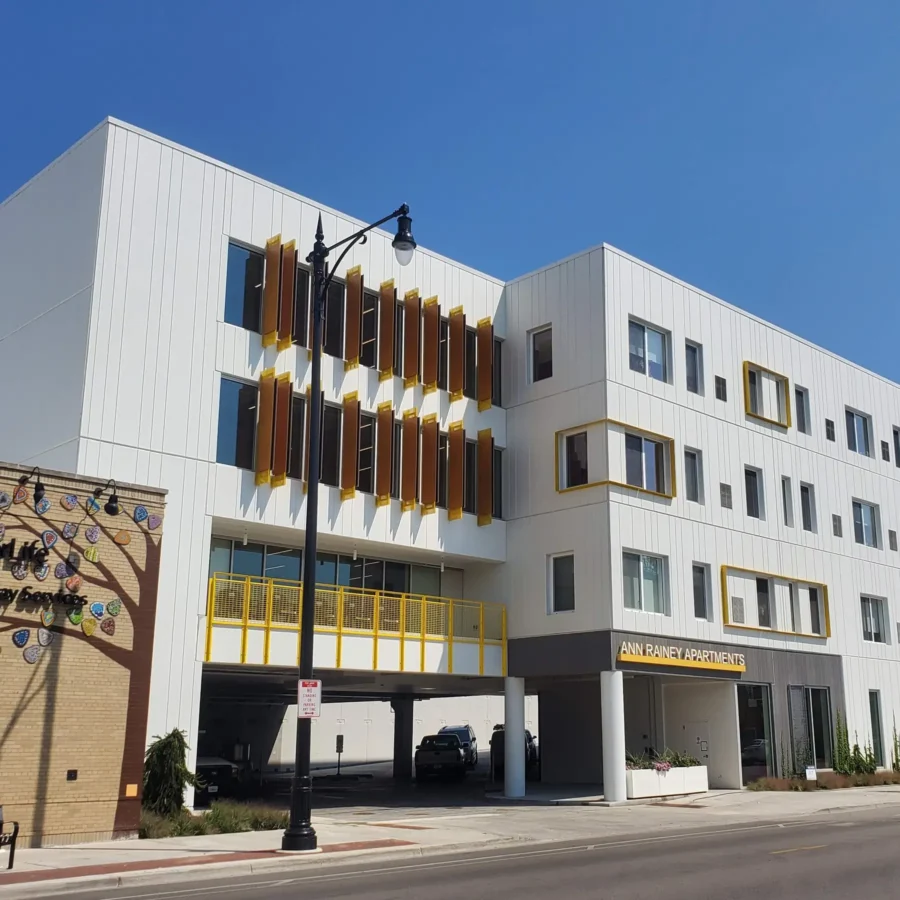 A modern, white multi-story apartment building with yellow accents and geometric window designs, named Ann Rainey Apartments, on a sunny day.