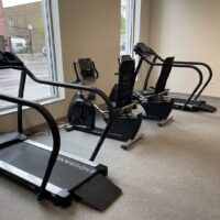 A small gym room with two treadmills and a recumbent exercise bike positioned near large windows facing outside.