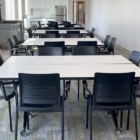 A modern meeting room with several rectangular tables arranged in rows and black chairs. A kitchen area with a refrigerator and sink is visible in the background.