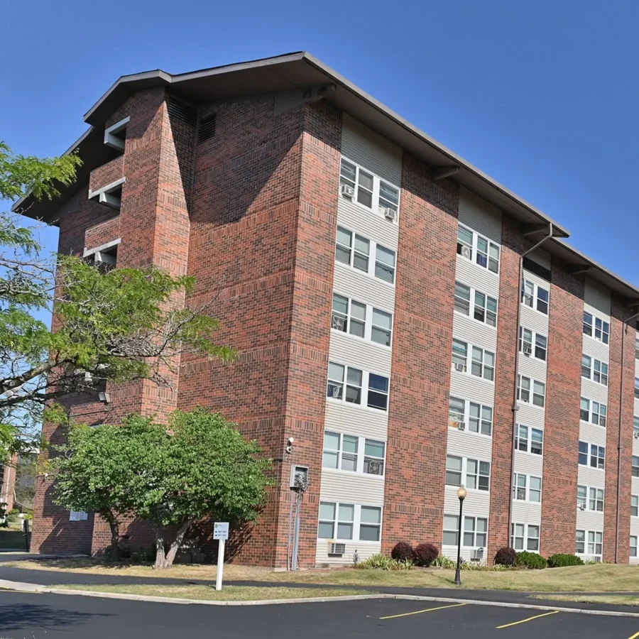A five-story brick apartment building with rows of windows, a sloped roof, and adjacent trees, photographed on a clear day.
