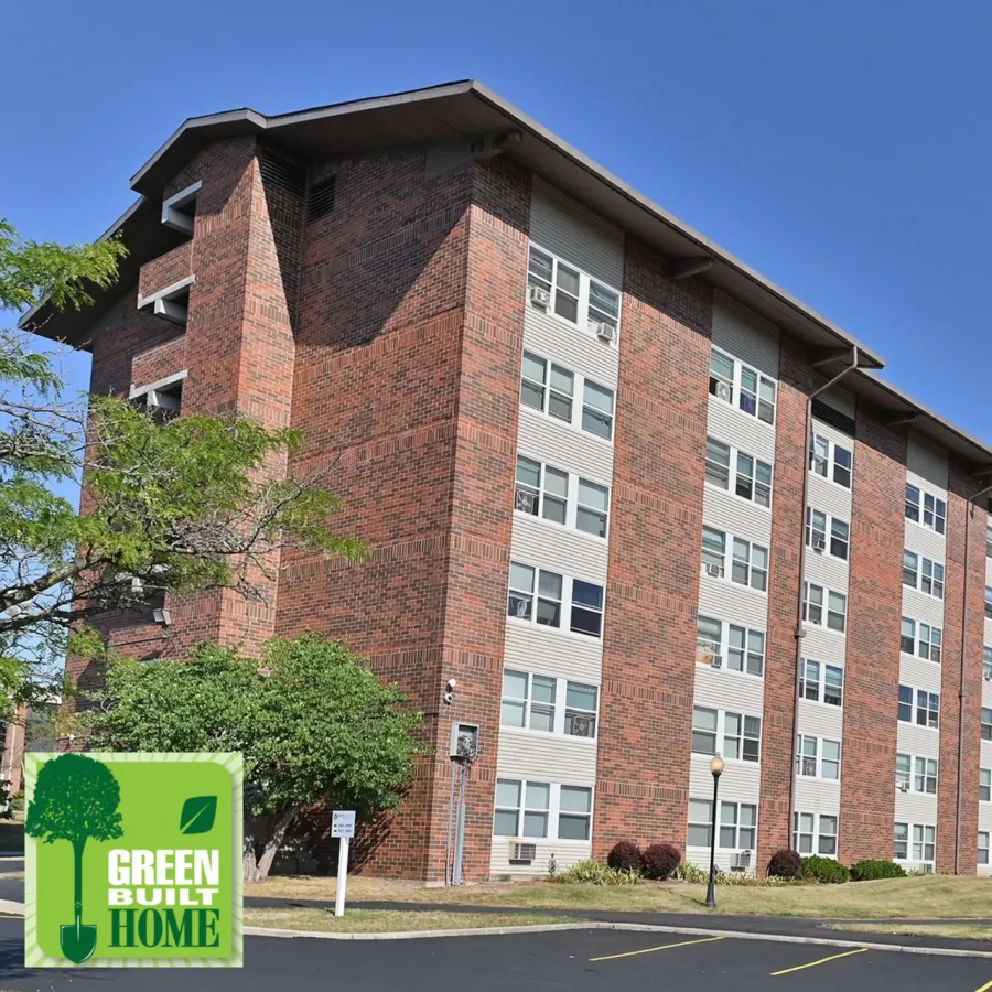 A five-story brick apartment building with white windows, a tree in front, and a “Green Built Home” logo in the lower left corner against a clear blue sky.