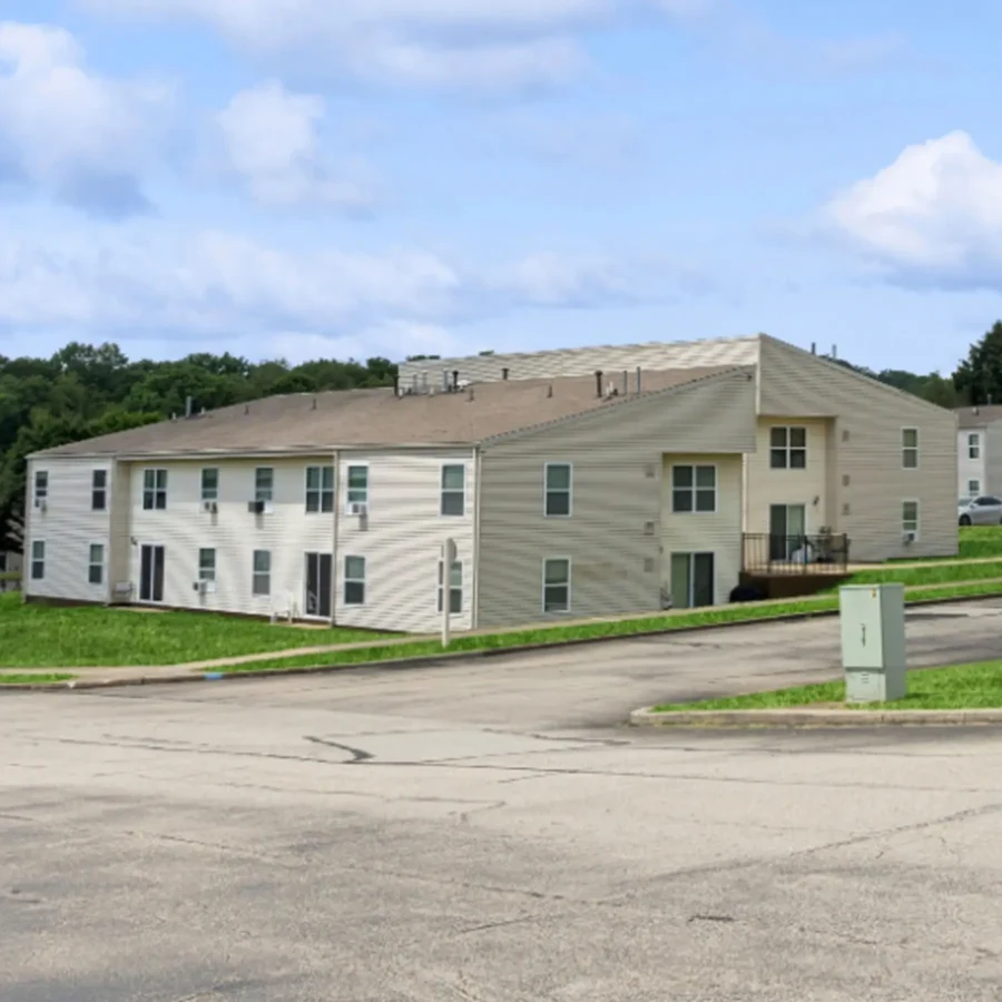 Two-story beige apartment building with multiple windows and balconies, situated on a grassy slope next to a paved road and a green utility box.