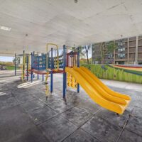 Covered playground area with colorful climbing structures, slides, and safety flooring, set against a mural-covered wall and a background of apartment buildings.