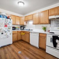A kitchen with wooden cabinets, white appliances, a refrigerator covered in magnets and papers, a dishwasher, and a stove; items and utensils are visible on the counters.